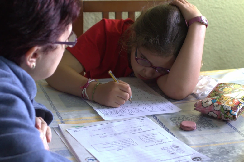 a girl sitting at a table with another person writing on paper