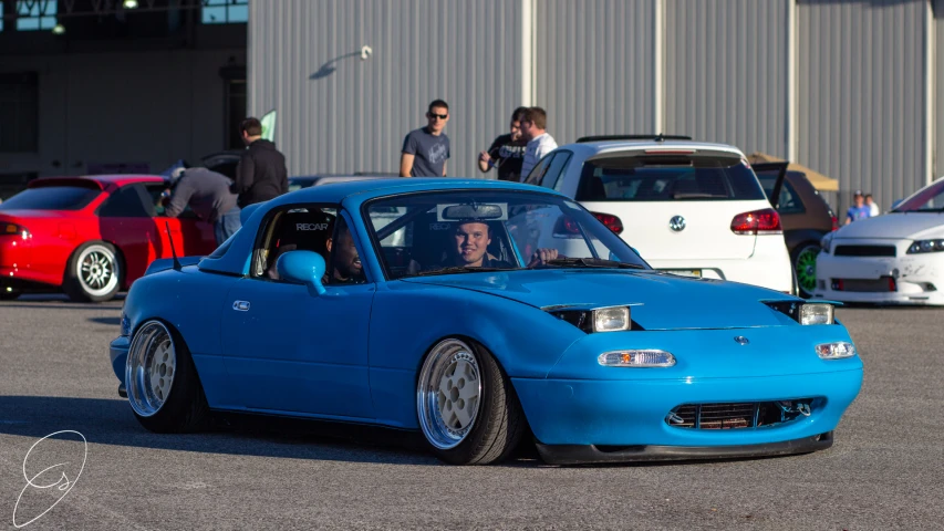 there is a blue colored car parked in front of a group of people