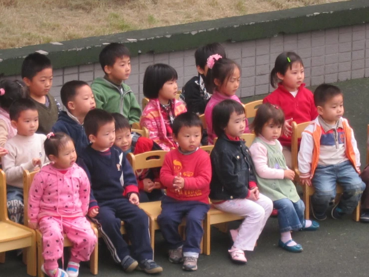 many children sit in wooden chairs outside in front of a wall