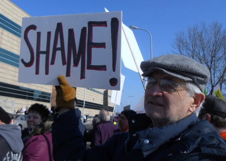 a man in hat and glasses holding up a sign with the word shame written on it