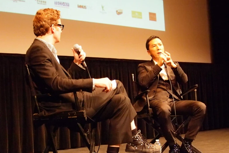 a pair of men sitting in chairs talking on stage