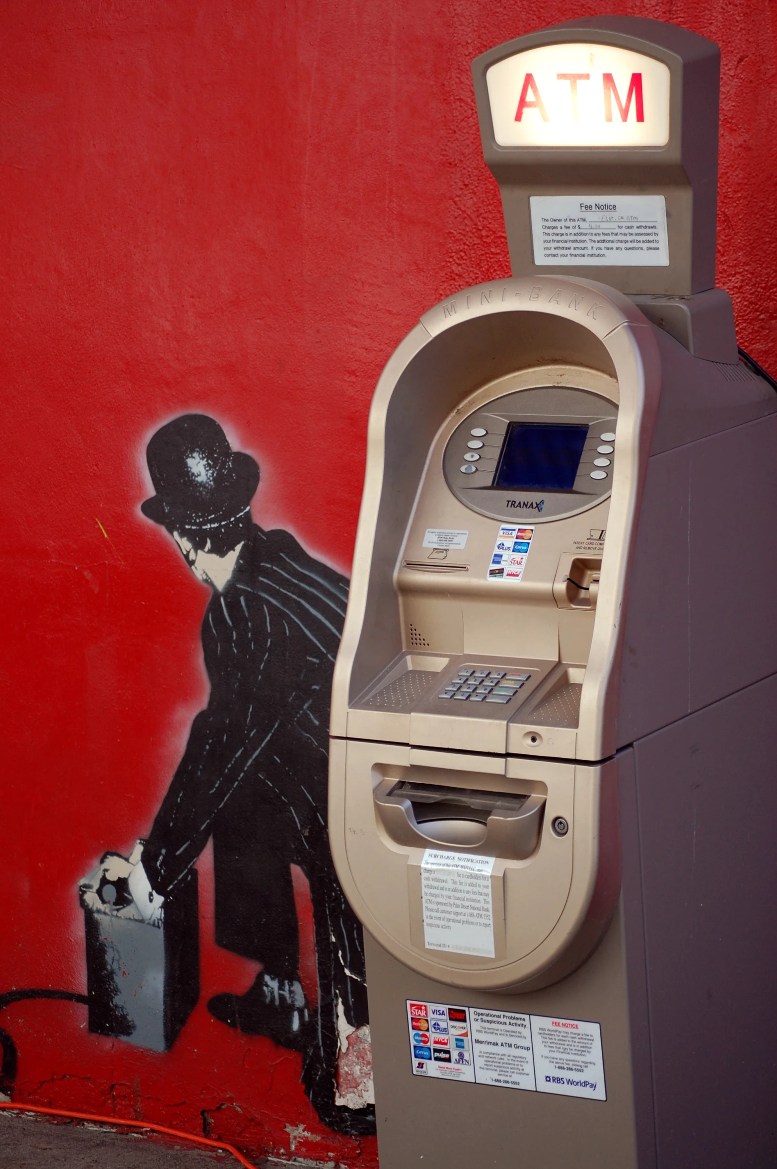 an atm in front of a red wall with a painting on it