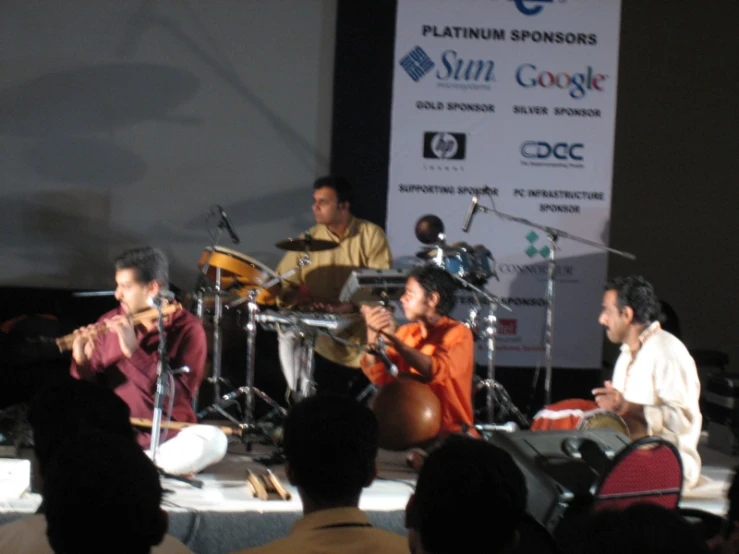 three men sitting on stage with instruments in front of them