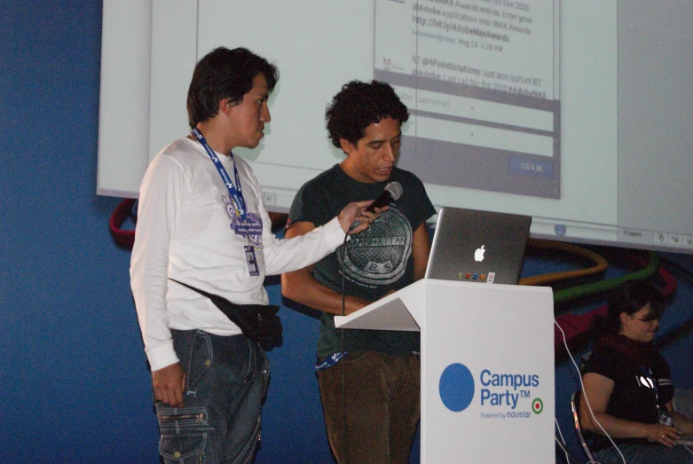 two men standing next to a computer in front of a projector screen