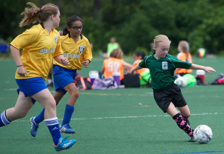 three children play a game of soccer with other players watching