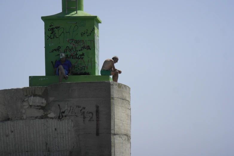 two people are sitting on the ledge of a green clock tower