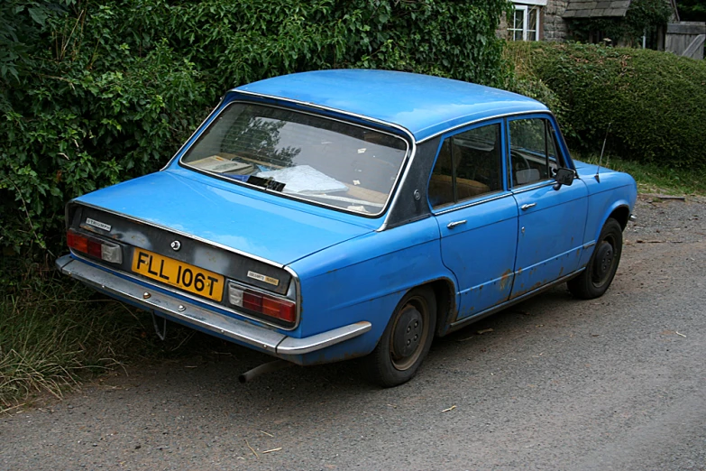 an old blue car parked on the street by a bush