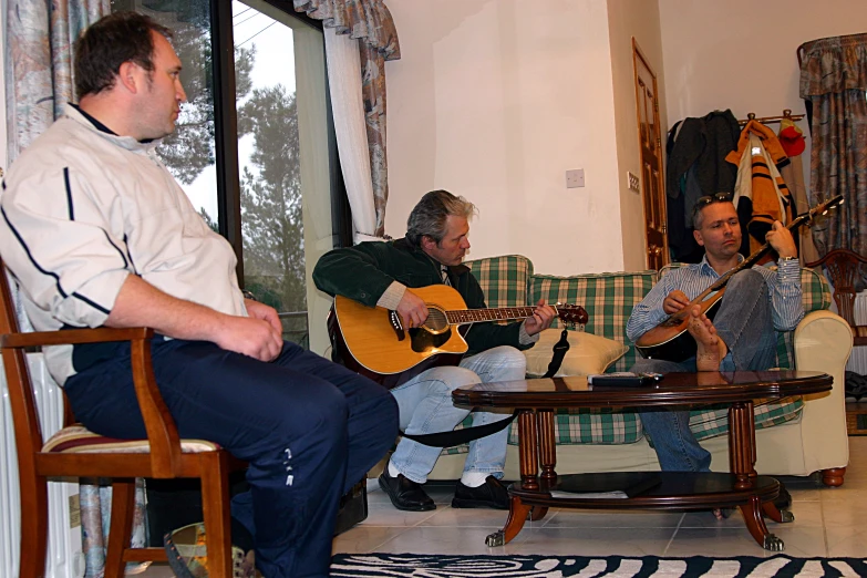 the men are sitting in a living room together playing guitar