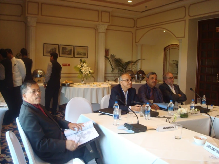 five men sitting around a table listening to some people speak