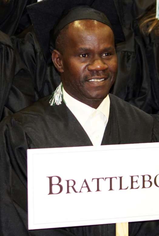 a smiling man in his graduation gown holding a sign