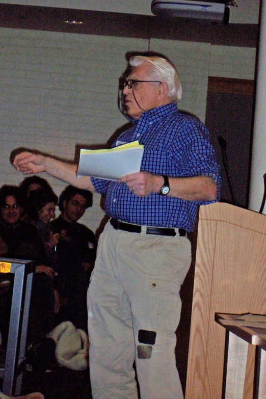a man wearing glasses and a blue checkered shirt is holding papers while speaking to a crowd