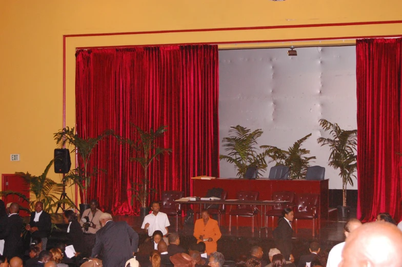 people sit and stand in front of a stage with red curtains