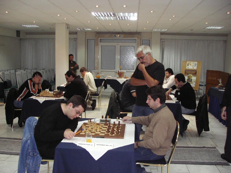 an older man plays chess with young men in a room full of tables