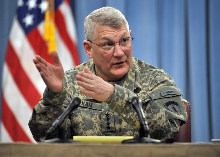 a man in uniform sits at a desk and holds his hand up