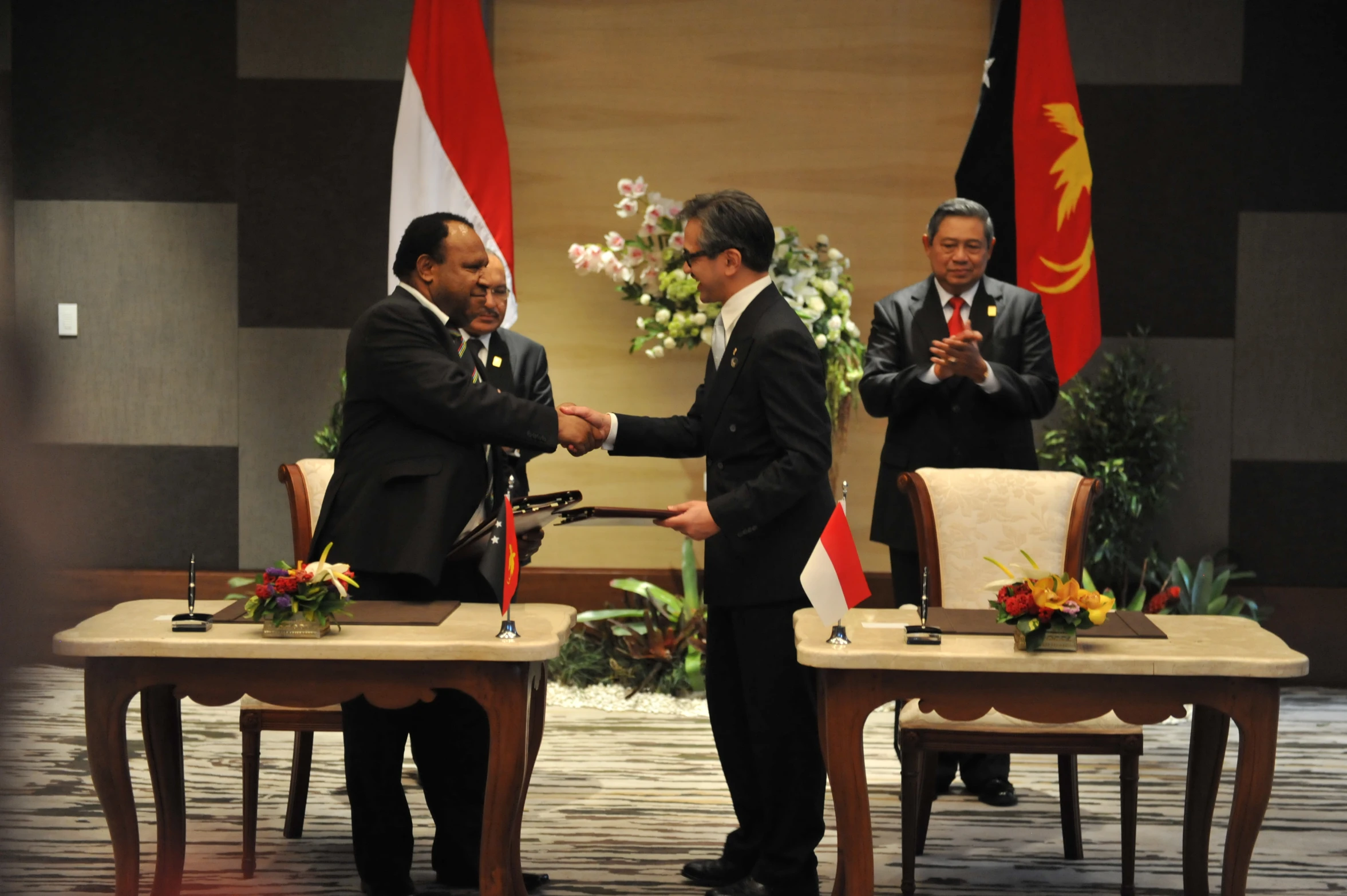 two men shake hands on the table while two flags fly around