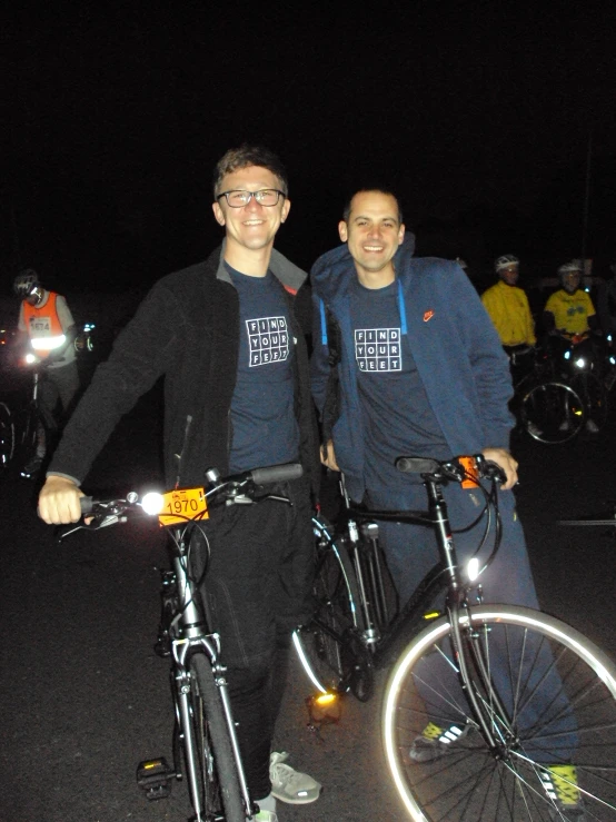two men standing next to bicycles while a dark background lights the street
