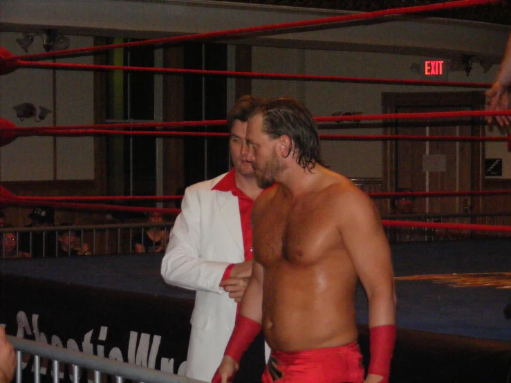 two men in red trunks and white jacket talking to each other in a wrestling ring