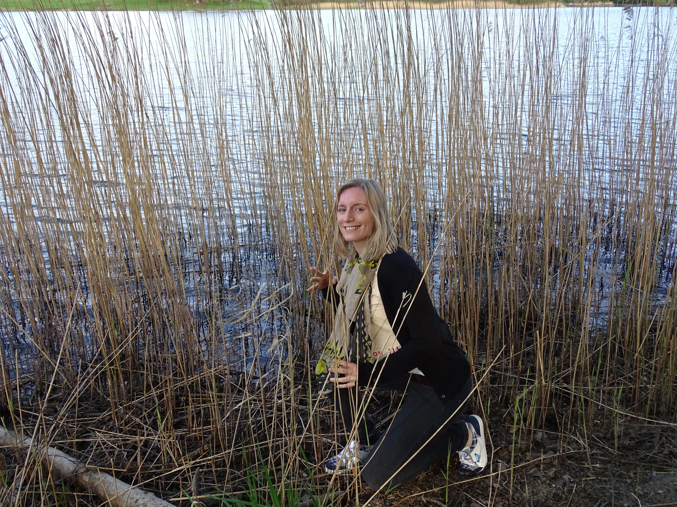 a smiling blonde woman crouched over plants by the water