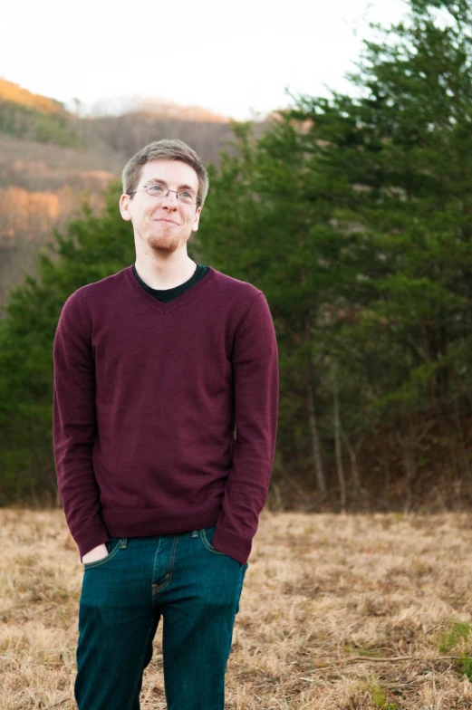 a man with glasses wearing blue jeans and a sweater standing in a field