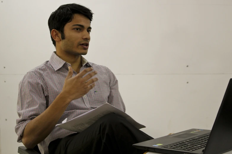 a man with a hand up sitting in front of a laptop computer