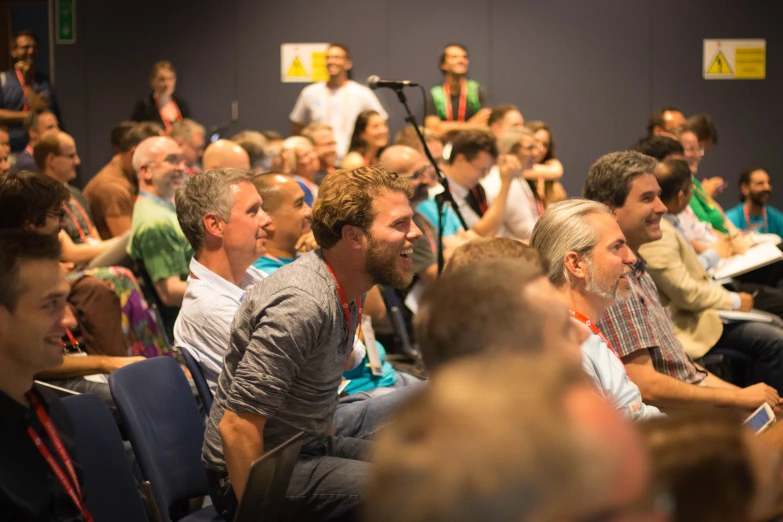 a large crowd sitting inside a room with microphones in the foreground