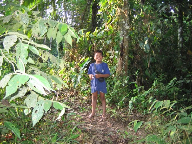 a boy stands in front of the camera in a lush, forested area