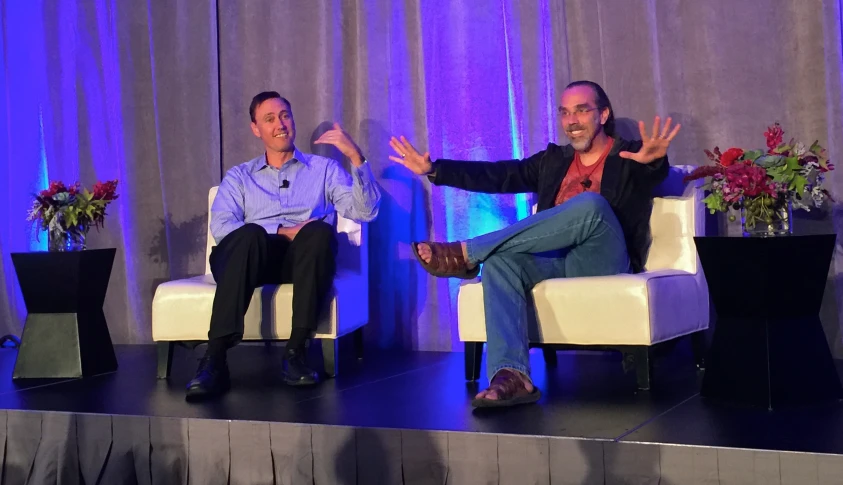 two men sit in chairs with their arms raised while a third man speaks