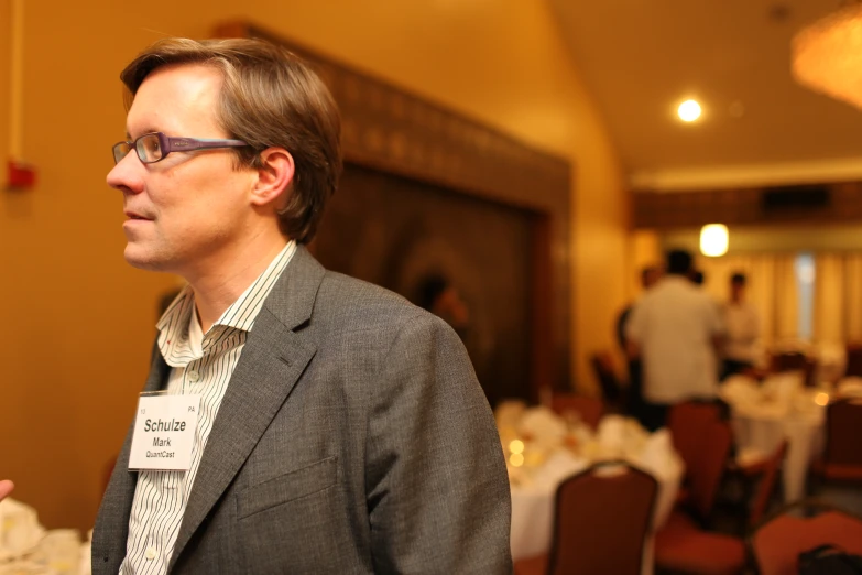 a man in suit with glasses standing next to a table filled with tables and chairs