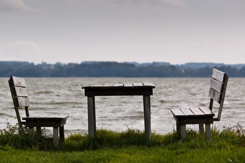 two benches and one chair with one missing sitting in grass next to water