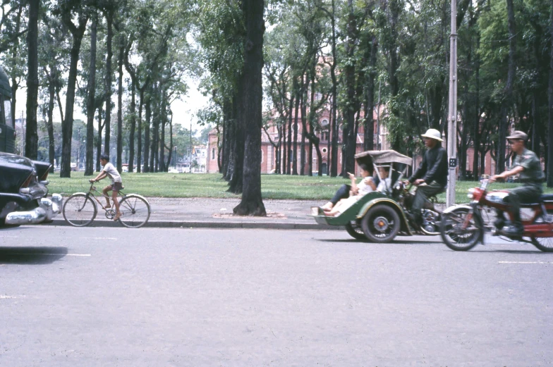 three people riding motorcycles, one has a passenger car, while the other a bike and a car are also carrying passengers