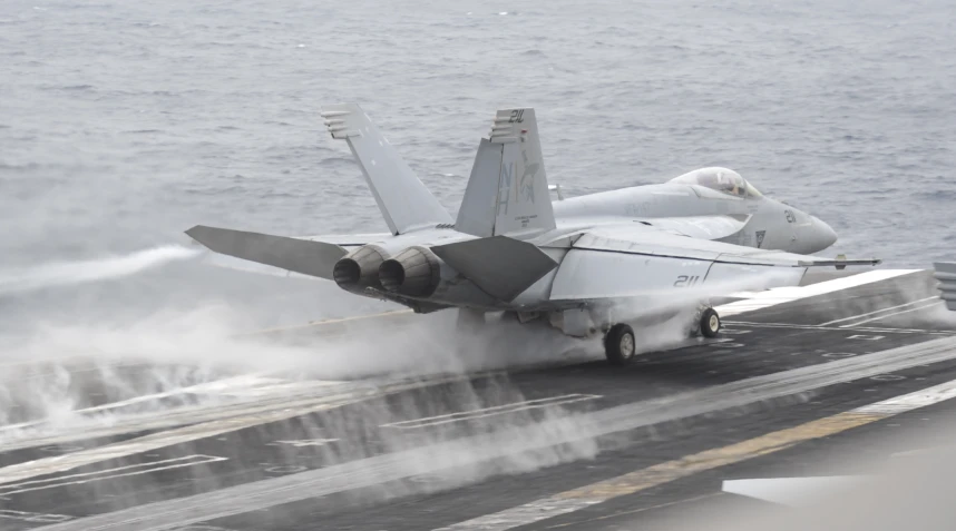 a navy fighter jet flying over the top of an aircraft carrier