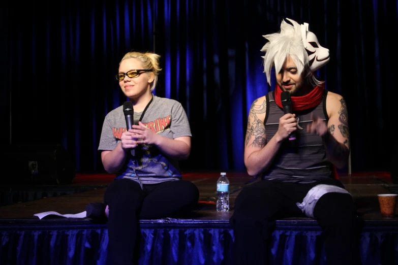 two women sit on stage talking while a man with tattooed face