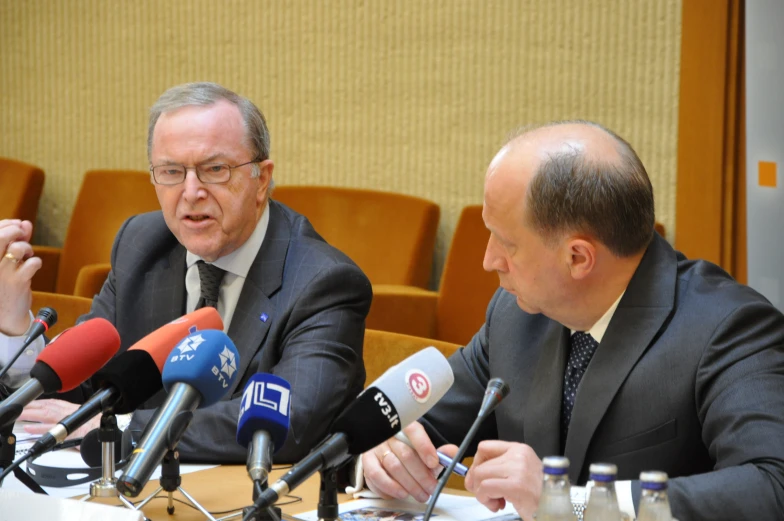 the man in a suit sits next to two other men sitting at a table with microphones on either side of him