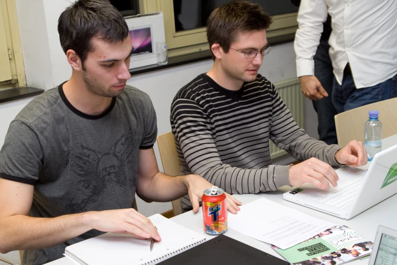 two young men sitting at a table working on laptops