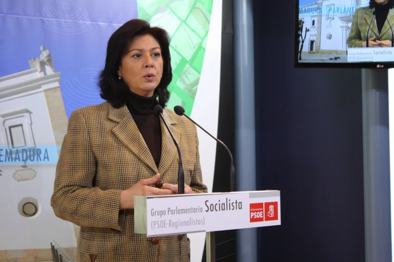 woman speaking at a podium near two televisions