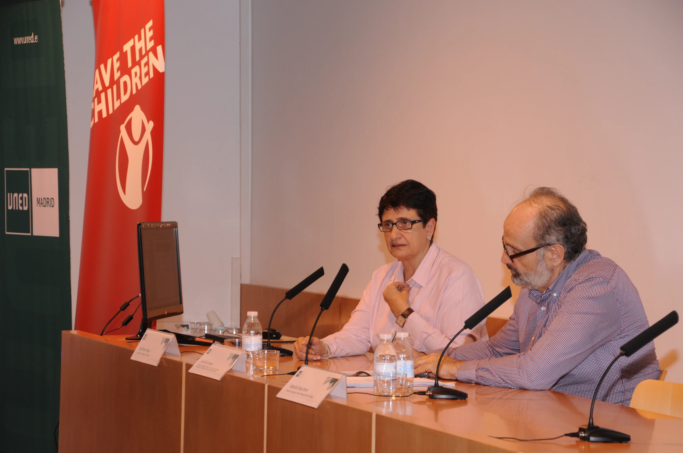 two men sit at a long table in front of microphones