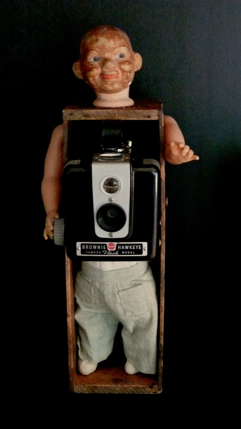 doll holding up an old camera in a crate