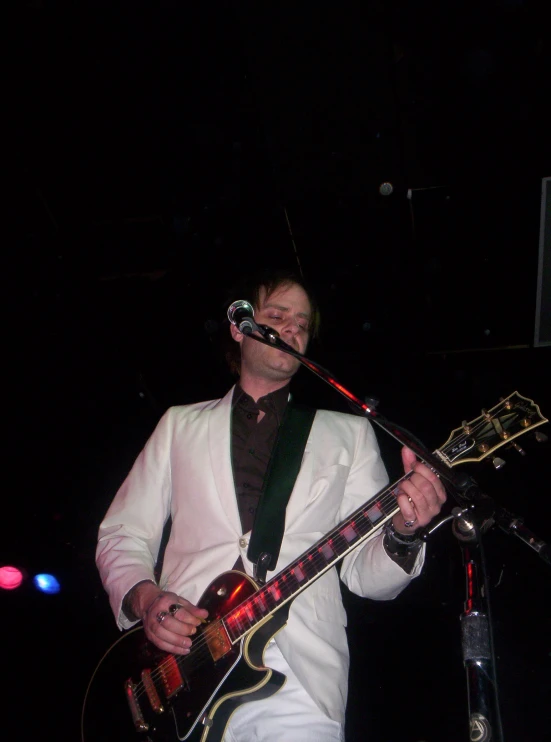 man singing into microphone while playing guitar with other people watching