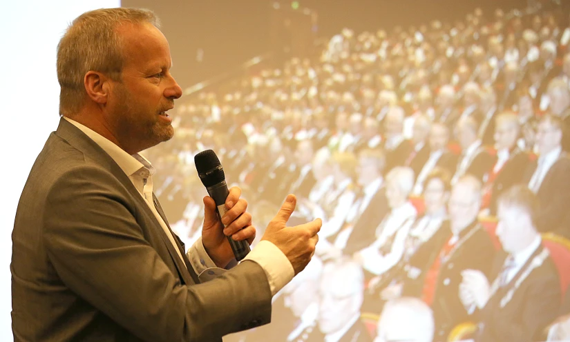 a man in suit talking into a microphone in front of a large screen