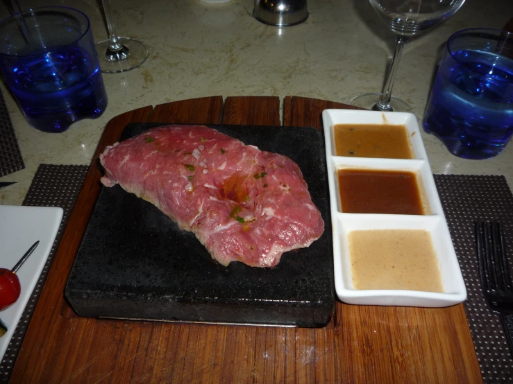 a large piece of meat on top of a  board next to two drinks and a spoon