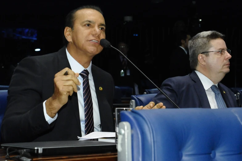 a man is speaking into microphone while a man with glasses on his head sits in front of him