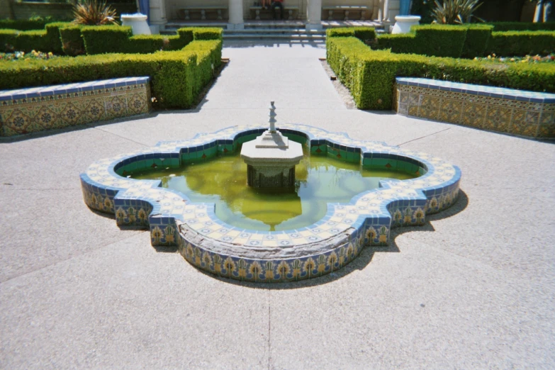 a fountain in the middle of a yard filled with hedges