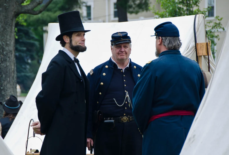 two old gentleman and a young man in hats talk near tents