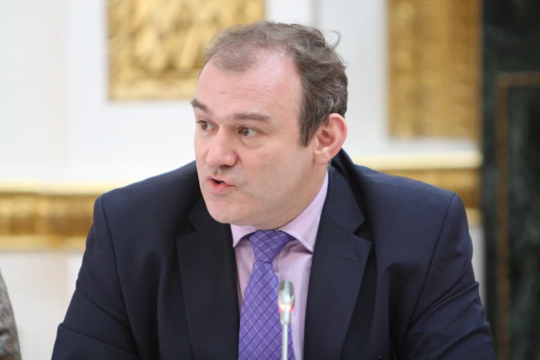 a man wearing a purple tie sitting at a table with microphone