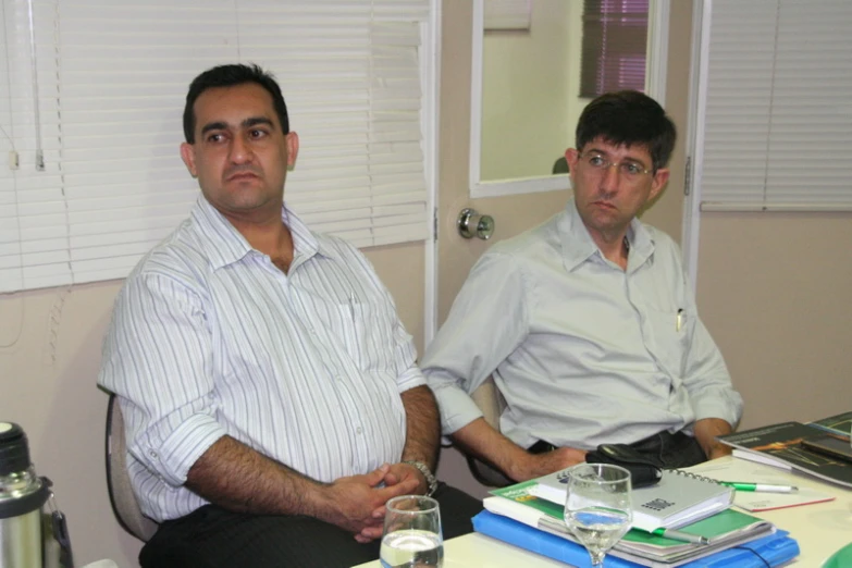 two men sitting at a desk, one with his hand on his hip