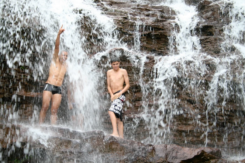 two boys standing next to a waterfall with water spewing over their heads