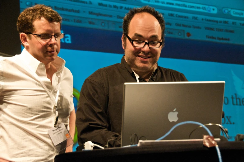 two men looking at an apple laptop in front of a screen