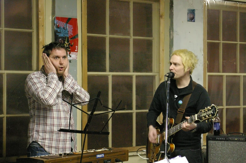 two men in a recording studio, one talking on the phone while the other plays a guitar