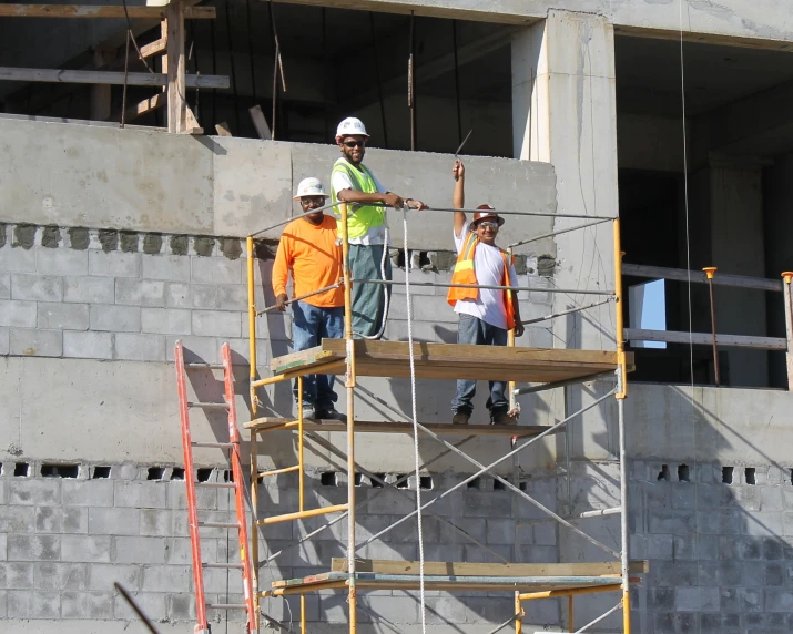 people wearing orange shirts stand on scaffolding with a brick building behind them