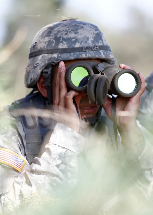 a soldier in fatigues wearing a helmet and a binch looking through binoculars